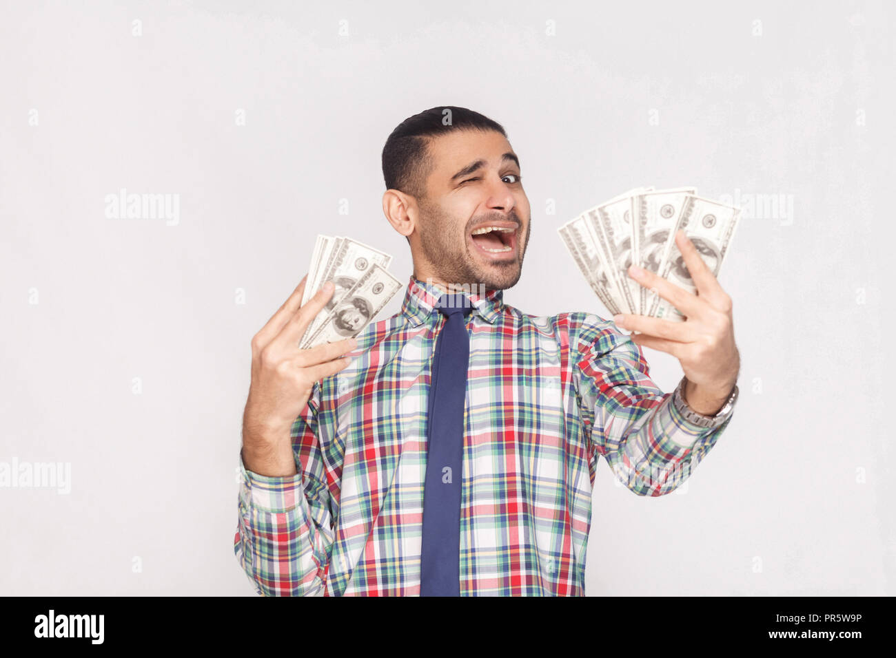 Joyeux Heureux beau jeune homme d'adultes en chemise à carreaux colorés avec cravate bleue debout, holding fan de dollars et en un clin d'avec la bouche ouverte. Banque D'Images Joyeux Heureux beau jeune homme d'adultes en chemise à carreaux colorés avec cravate bleue debout, holding fan de dollars et en un clin d'avec la bouche ouverte. Banque D'Images