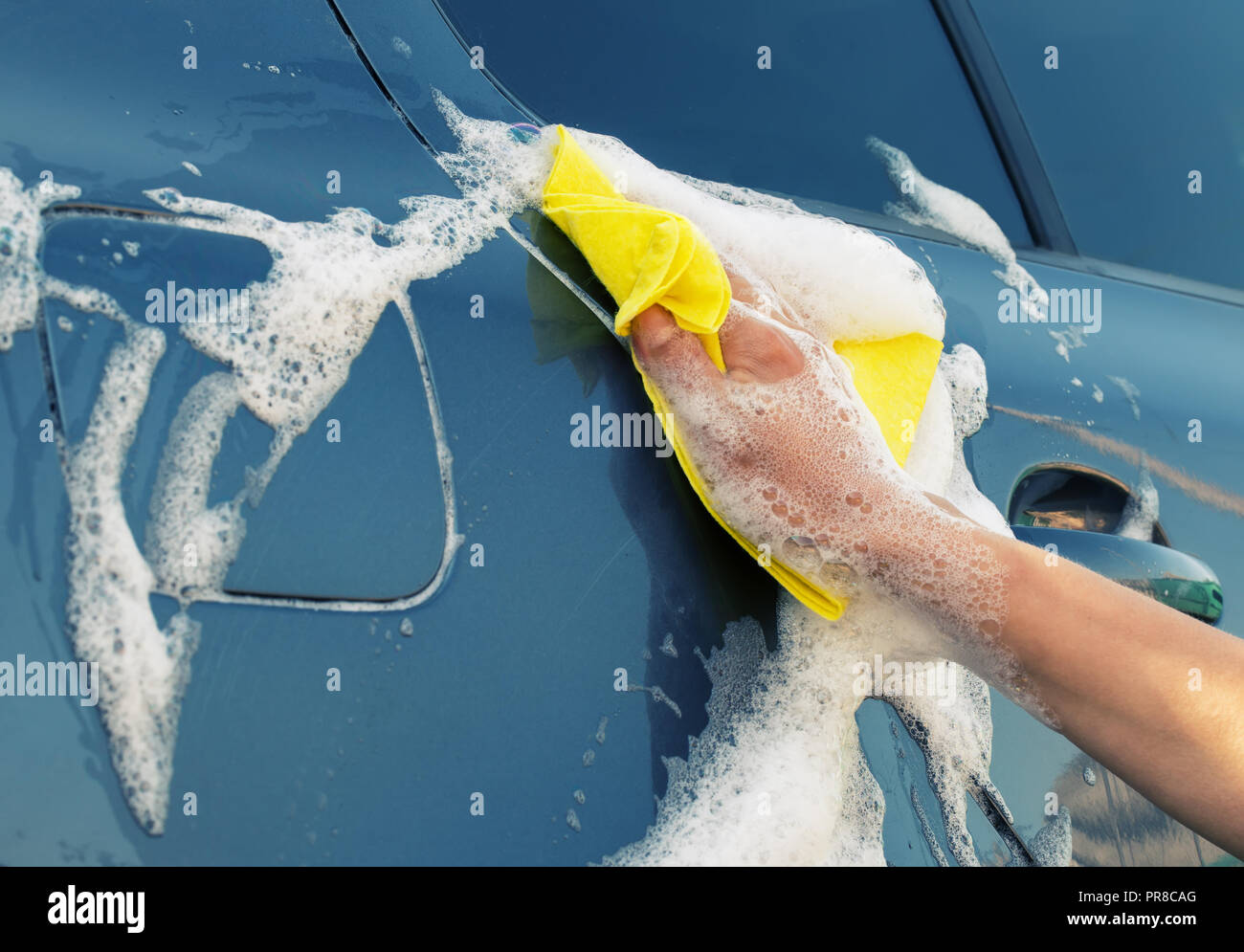 Laver la voiture grise avec un chiffon jaune en mousse savonneuse. A woman's hand wipes le côté de la voiture Banque D'Images