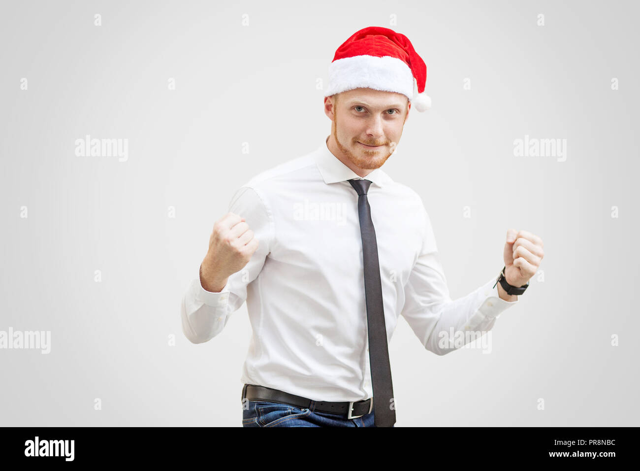 Happy handsome businessman en chemise blanche, cravate noire et rouge nouvelle année hat, looking at camera avec sourire et celebrting avec bras levés. Piscine studio Banque D'Images