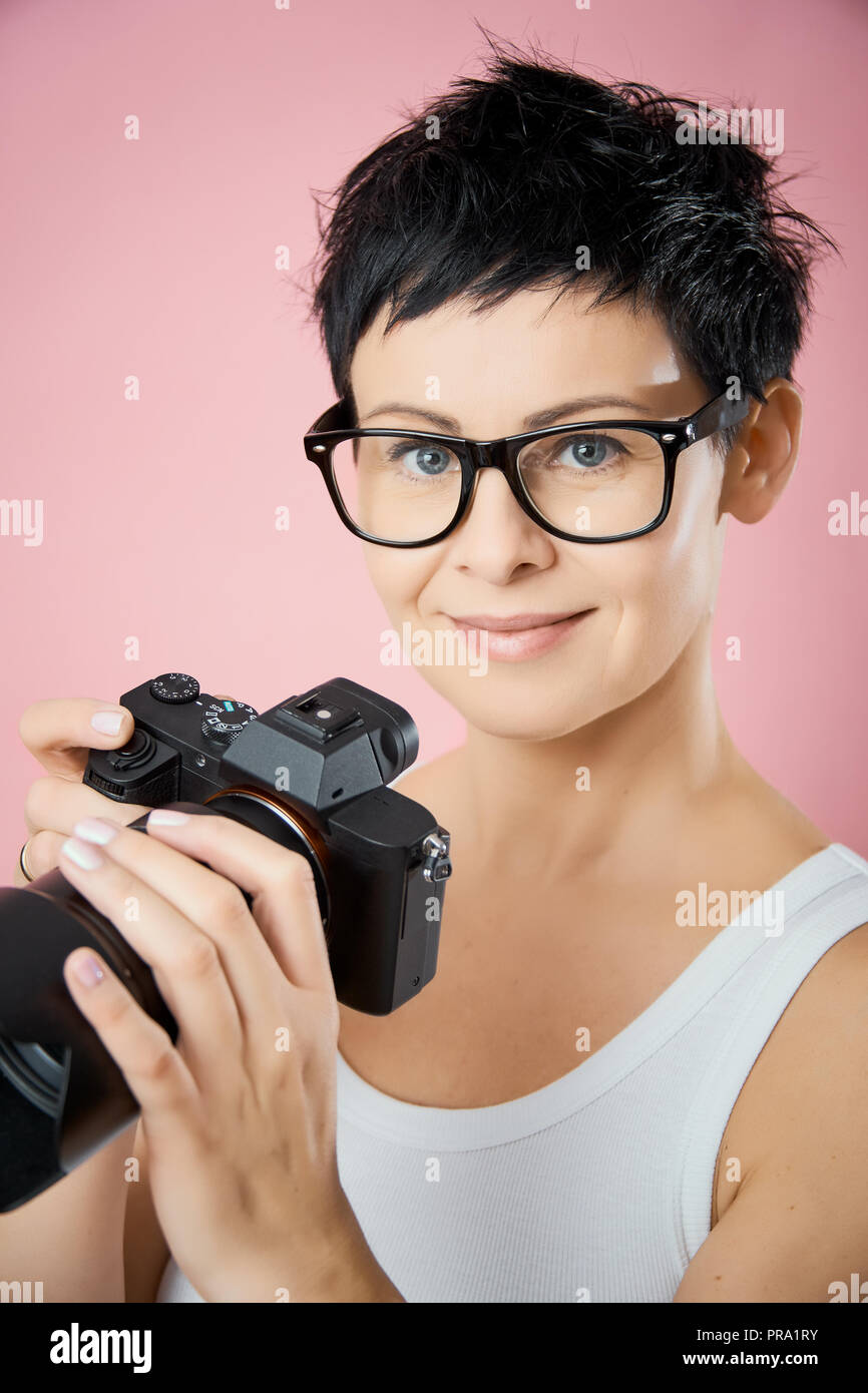 Belle femme à lunettes et aux cheveux courts avec un appareil photo en main  pose sur un fond rose Photo Stock - Alamy