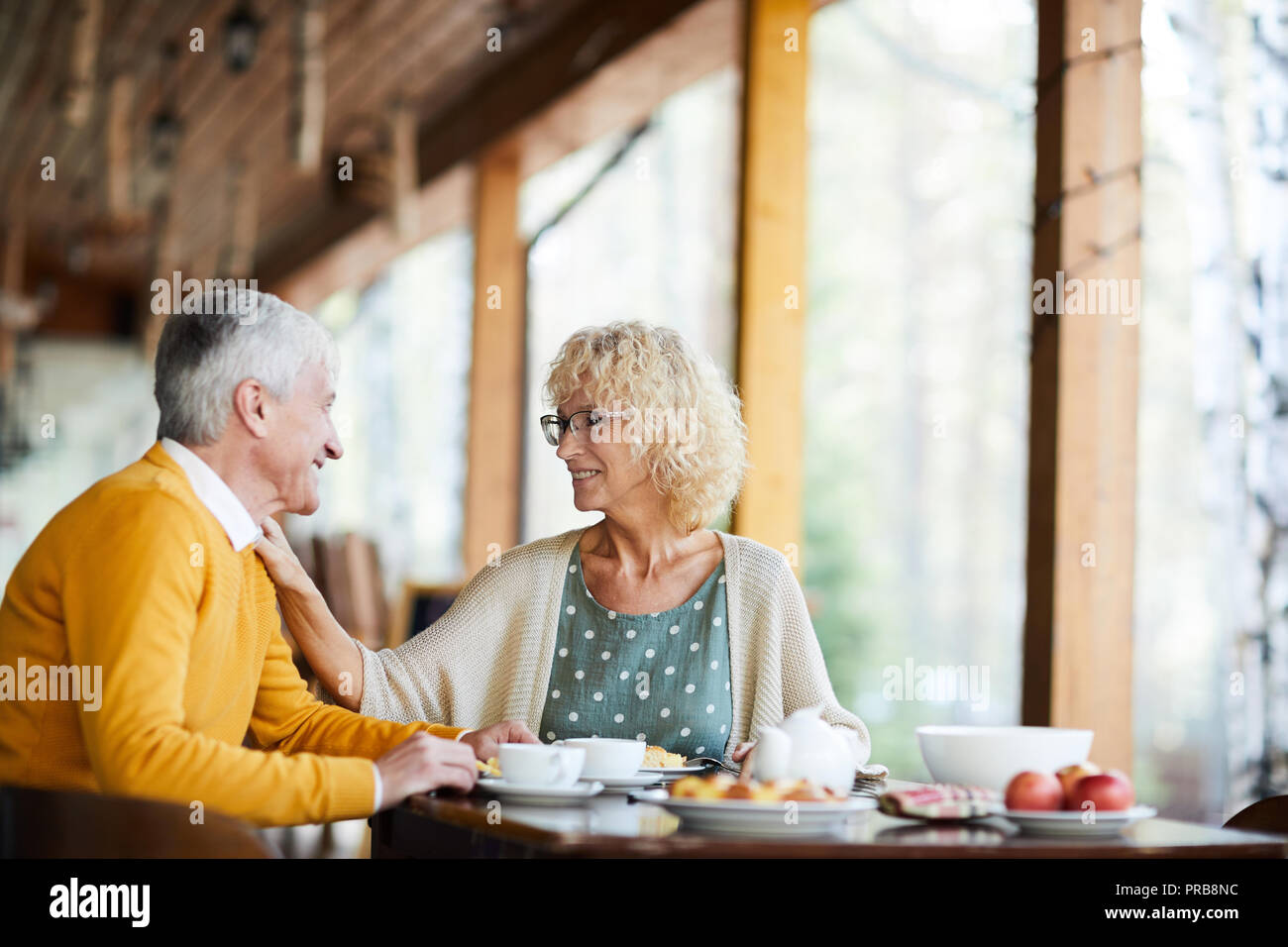 Happy senior femme avec les cheveux bouclés portant des lunettes de toucher l'épaule du mari et le soutenir pendant qu'ils parlent au petit-déjeuner sur le porche Banque D'Images