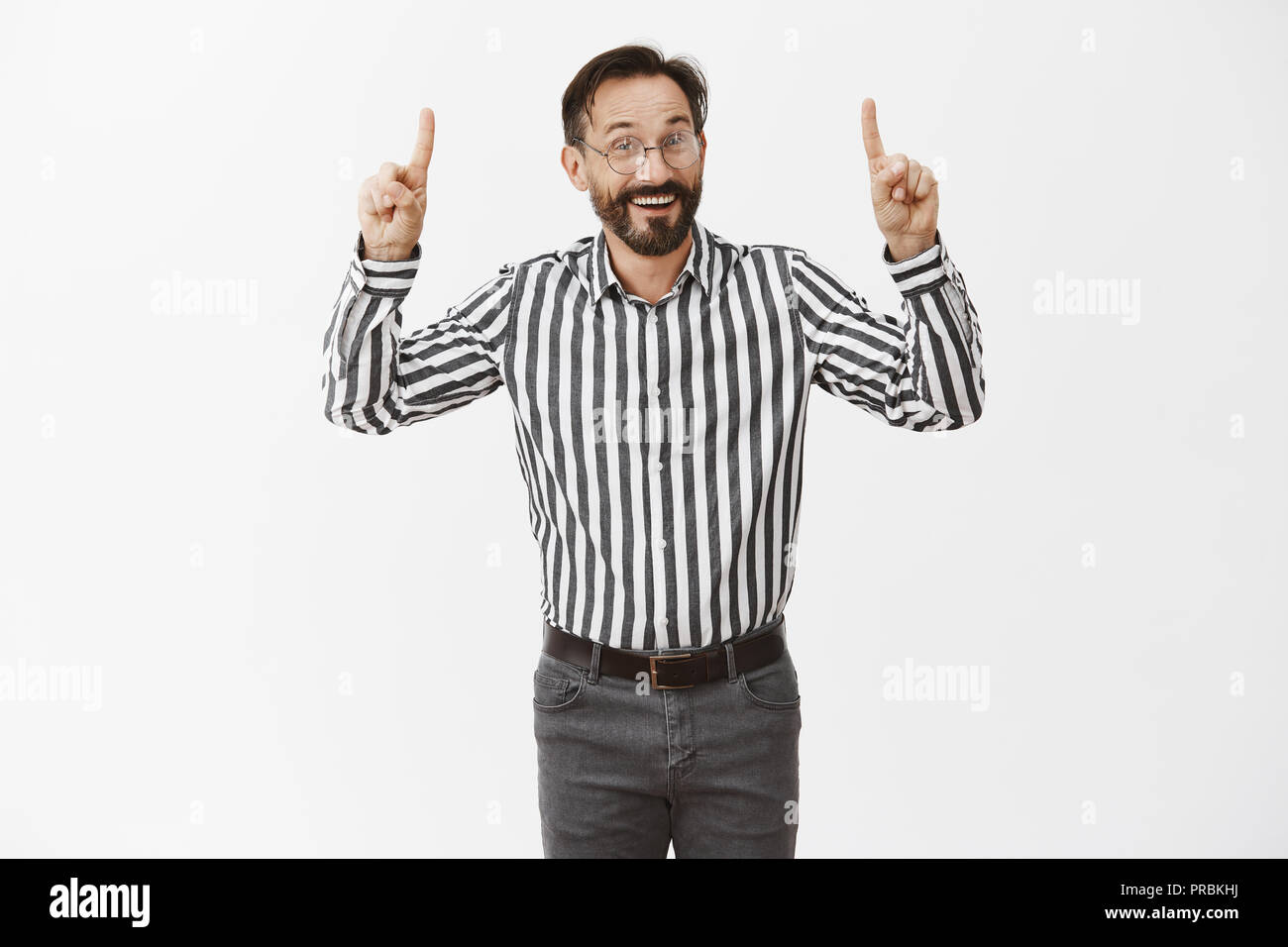 Regardez en haut cher invité. L'homme charmant et hospitalier avec barbe, dans les verres et chemise rayée, pointant vers le haut avec les doigts et tout sourire tandis que la publicité ou la promotion de sa campagne Banque D'Images