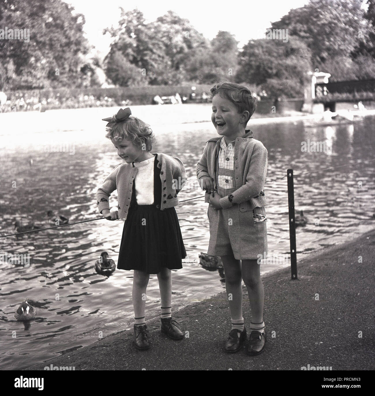 Années 1950, historique, un jeune garçon et fille debout par un grand étang ou lac de plaisance, avec le garçon ayant trouvé quelque chose de très amusant, rire, tandis que sa sœur, ratherly timidement, examine les canards, England, UK. Banque D'Images