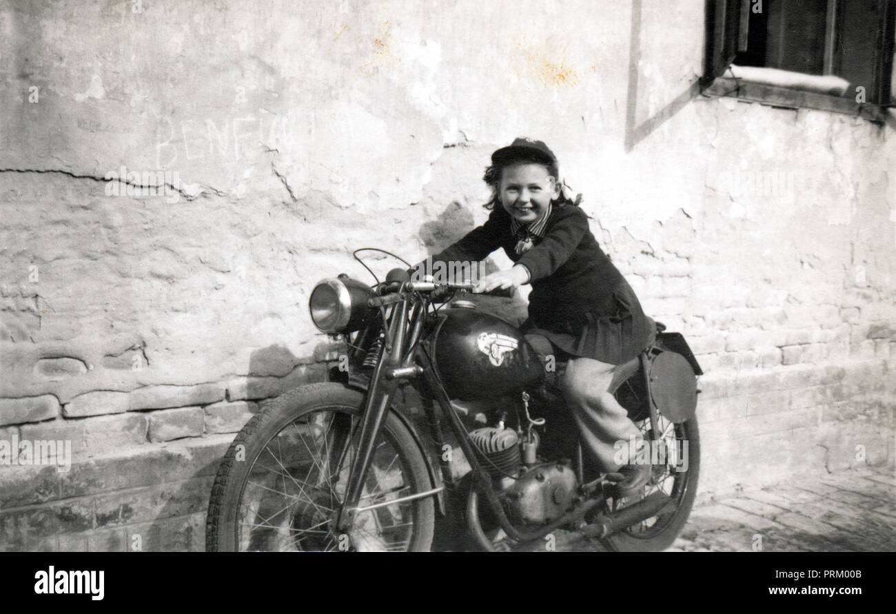 Jeune fille pose pour photo sur une moto vétéran sur la chaussée à côté d'un mur de briques érodées vers 1948 Banque D'Images