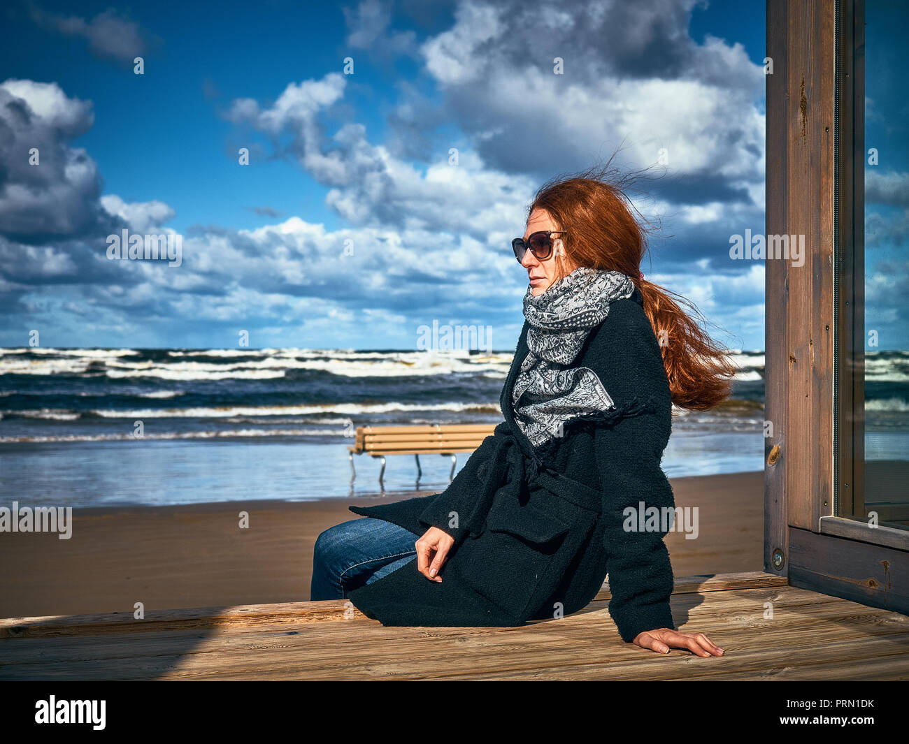 Fille aux cheveux roux admire la vue de la mer assis sur le morceau de marbre de la terrasse ouverte. La jeune fille est vêtue d'un manteau court noir automne et bleu Banque D'Images