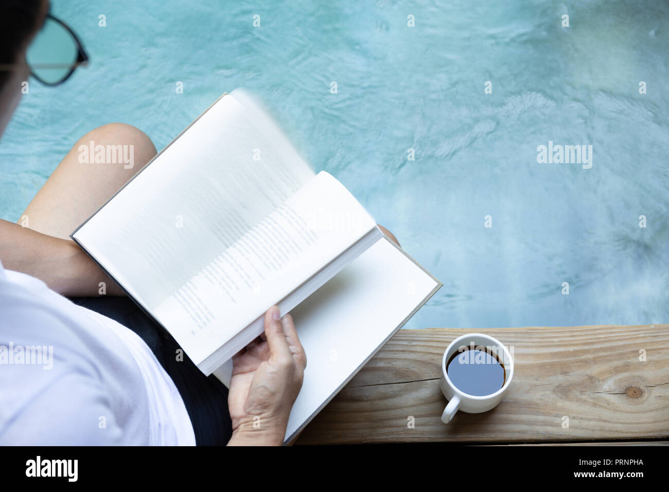 Man reading book avec tasse de café sur un plancher en bois à côté d'une piscine extérieure Banque D'Images