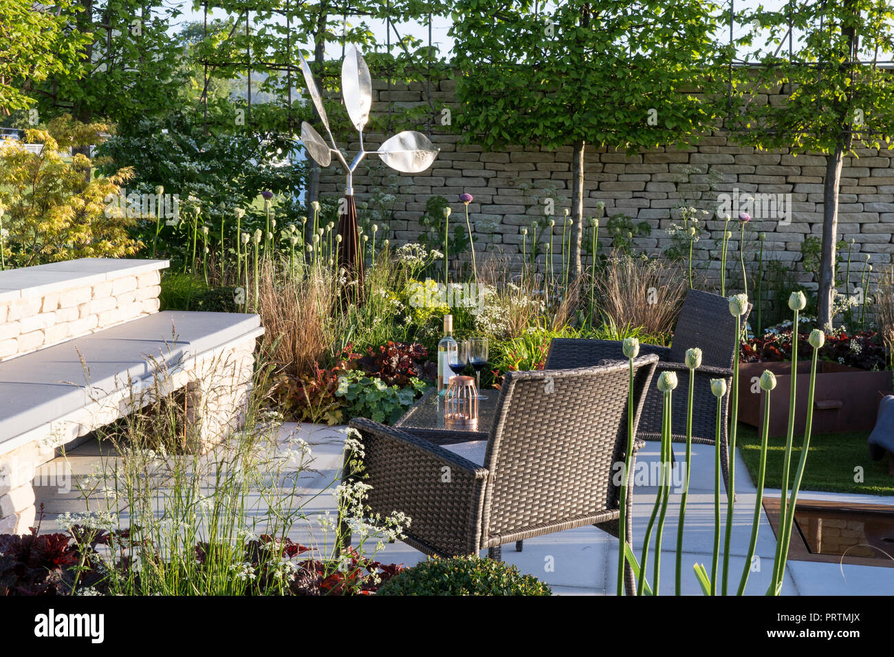 Patio en pierre avec des meubles de jardin en rotin, banc en pierre, mur en pierre de Cotswold à sec, pleached Carpinus betulus arbres, graminées, Allium, sculpture en acier, sp Banque D'Images