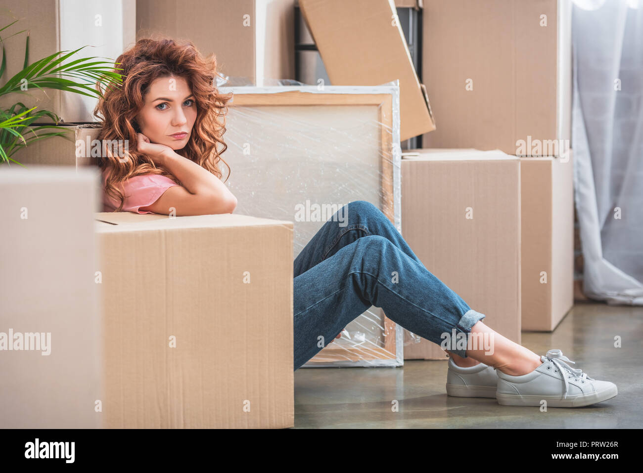 Jolie femme avec les cheveux rouges assis sur le plancher près de boîtes de carton à nouveau la maison et looking at camera Banque D'Images