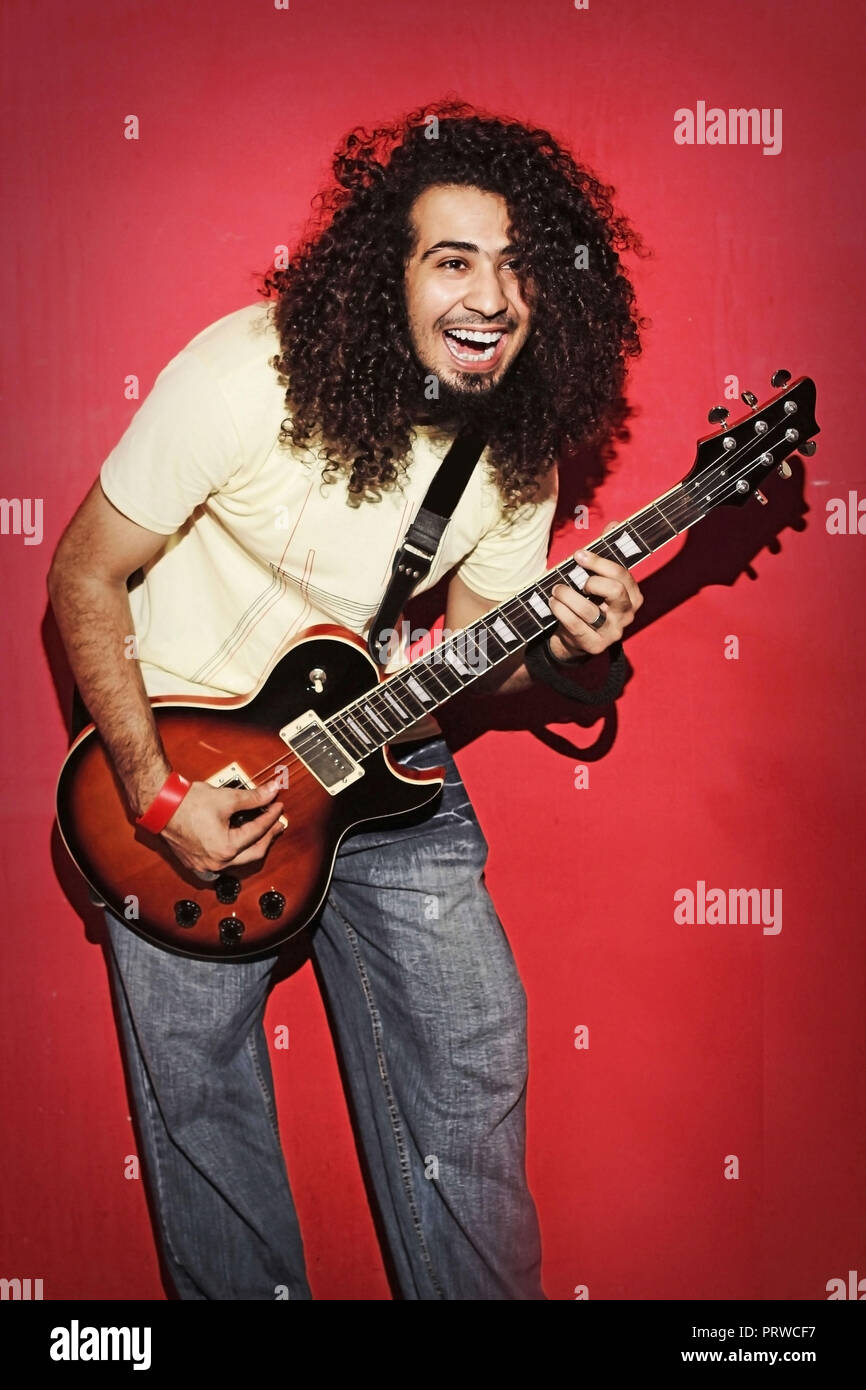 Gros plan d'un bel homme passionné expressif jeune musicien de rock brunette cool avec des cheveux longs jouant de la guitare électrique contre le rouge Banque D'Images