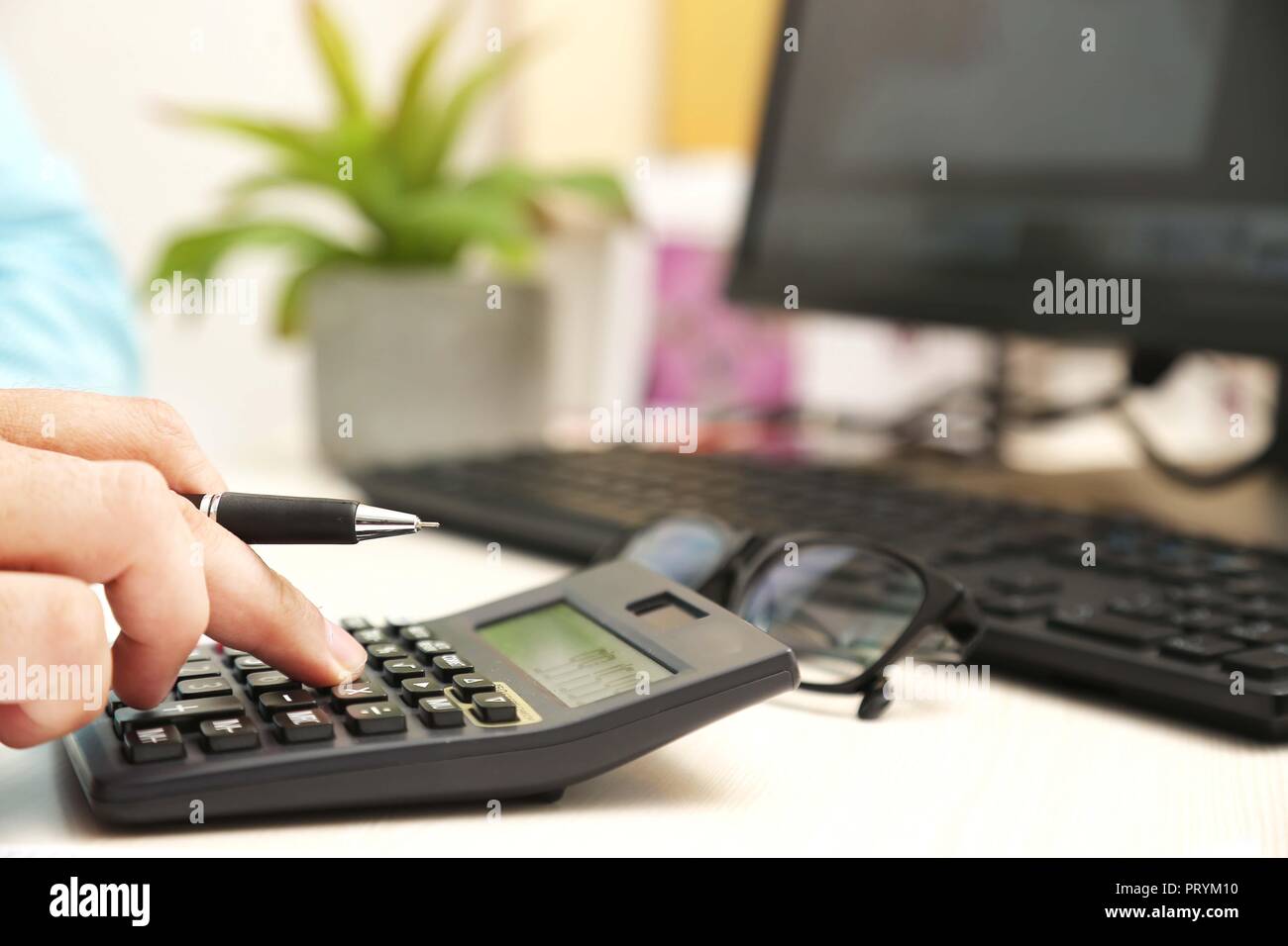 L'homme est l'utilisation de calculatrice avec stylo à la main. Photo de l'ordinateur, clavier, pot de fleurs et des verres sur la table. Banque D'Images