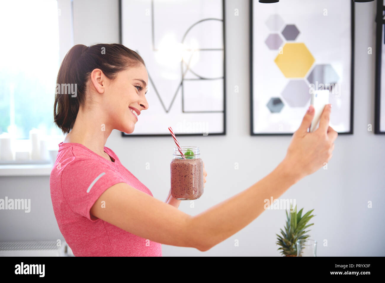 Jeune femme avec des smoothies avec selfies smartphone dans la cuisine Banque D'Images