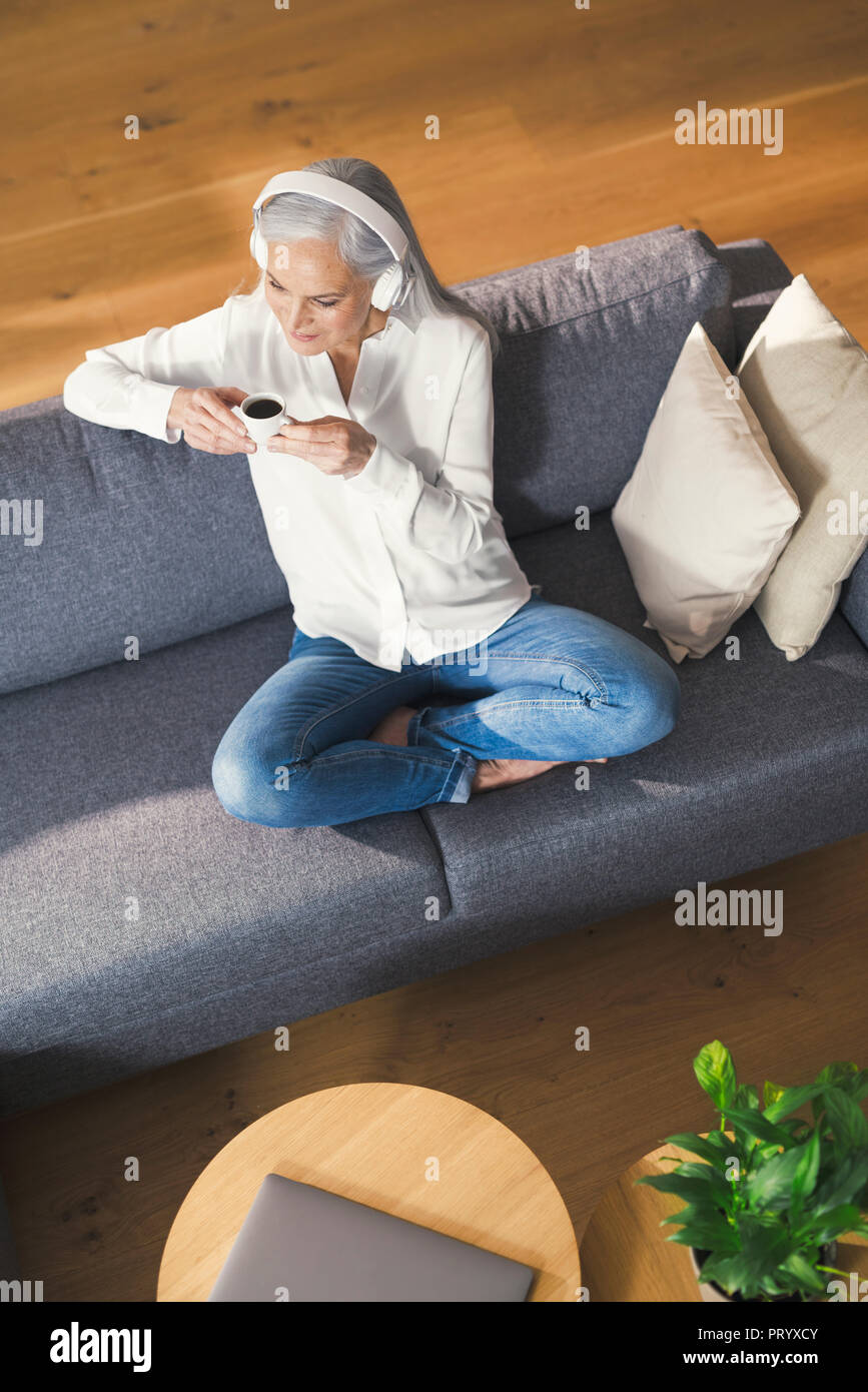 Senior woman sitting on couch, écouter une musique ning boire du café Banque D'Images
