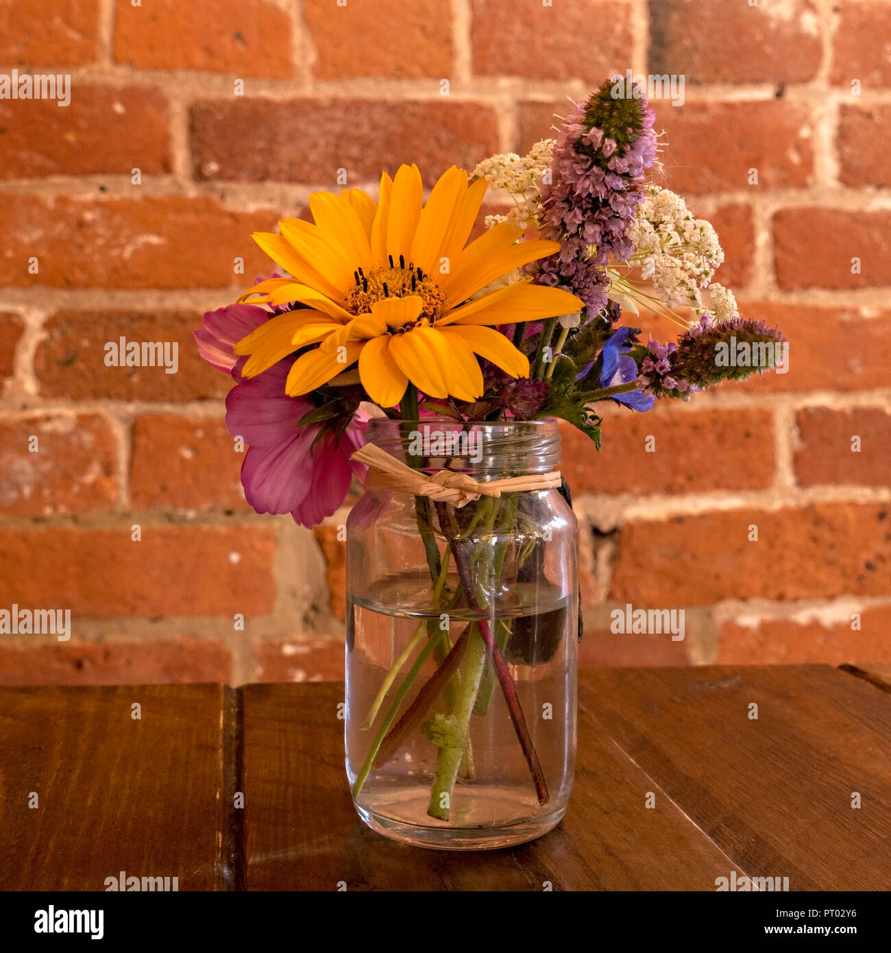 Jolie décoration de table - petit pot de confiture en verre clair avec posy arrangement de fleurs sur table en bois avec mur de brique rouge derrière, UK Banque D'Images