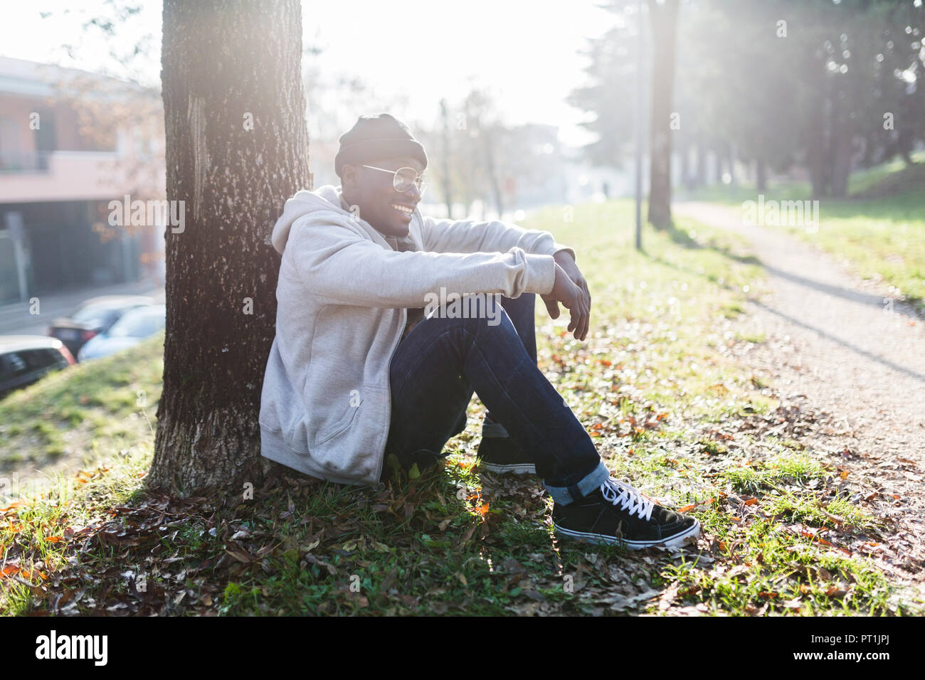 Laughing young man sitting in park, leaning on tree trunk Banque D'Images