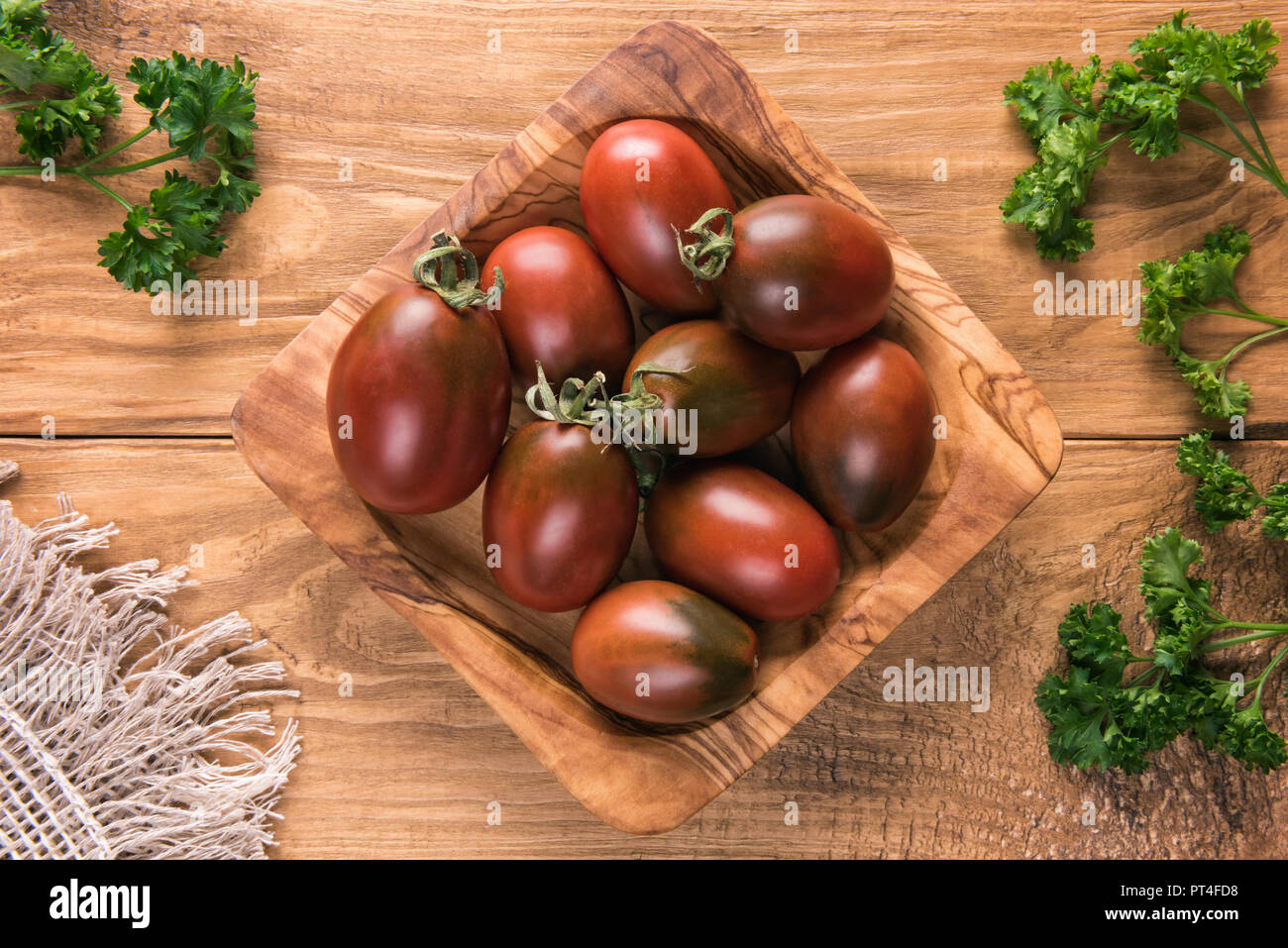 Vue de dessus sur les petites rouge foncé Ovale ou "noir" les tomates dans le bol en bois naturel carré en bois avec surface texturée table vert les feuilles de persil. S Banque D'Images