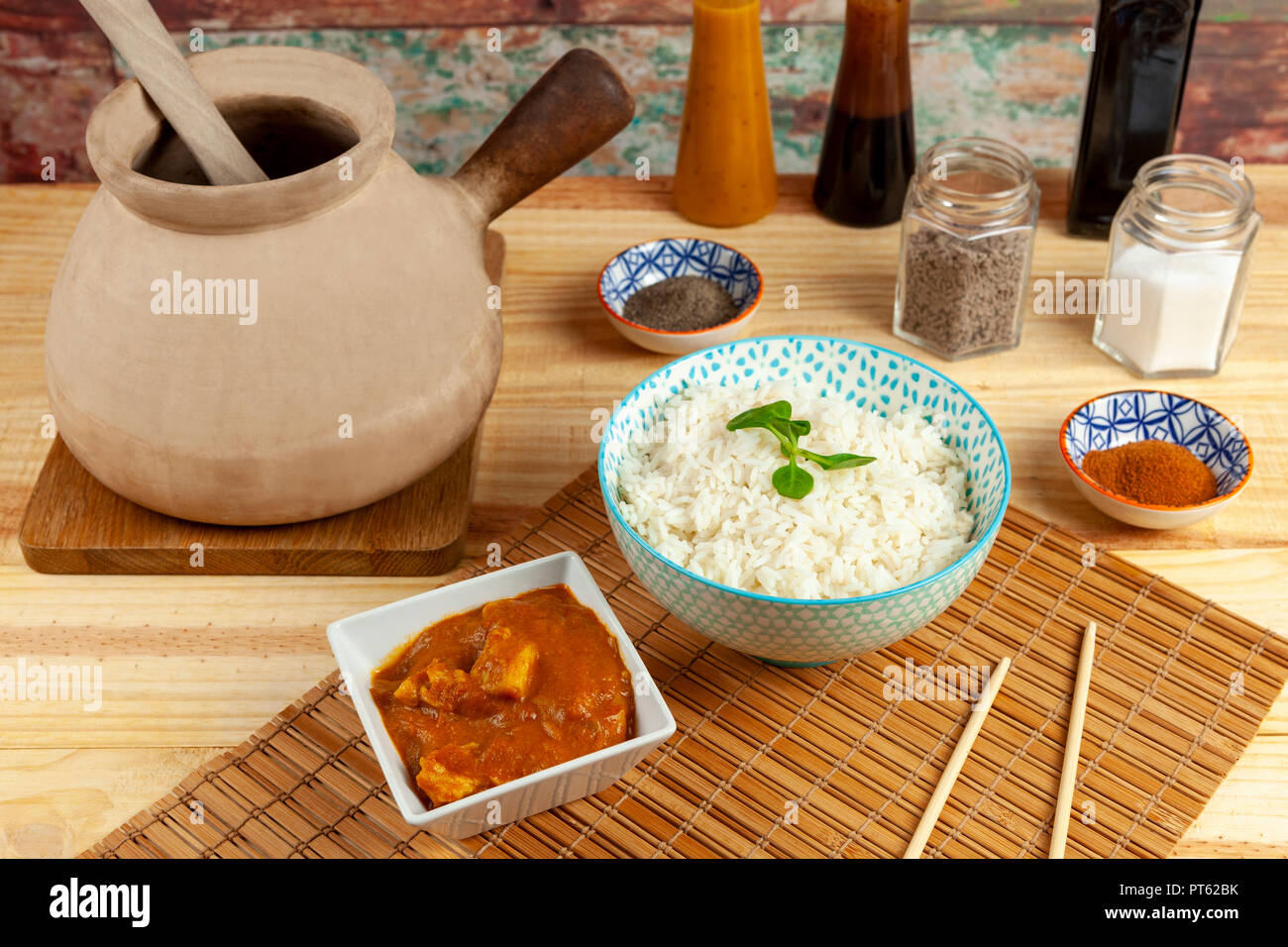 Bol de riz et d'une partie de poulet au curry disposés sur un tapis de bambou le long d'une marmite traditionnelle chinoise et condiments sur une table en bois Banque D'Images