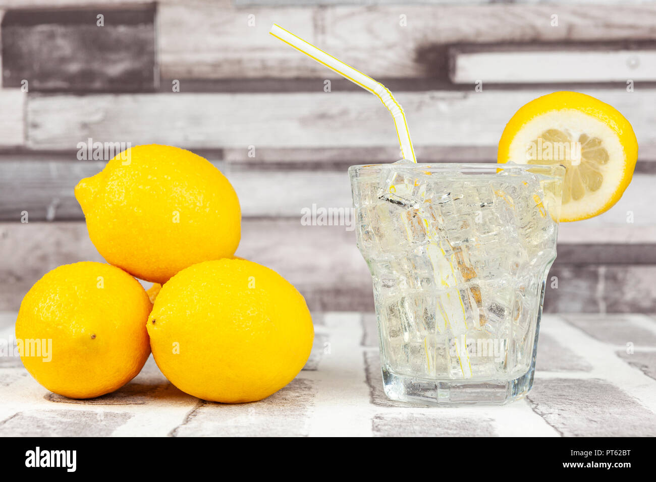 Verre de l'eau de citron glacé avec une paille et une tranche de citron de côté un tas de citrons entiers frais mûrs Banque D'Images