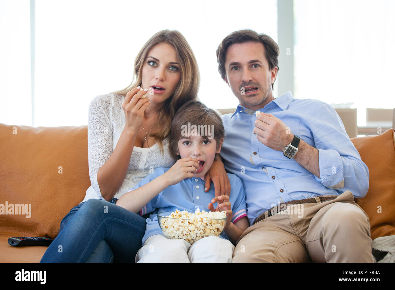 Beaux jeunes parents et leurs fils sont devant la télévision, eating popcorn sitting on sofa at home Banque D'Images