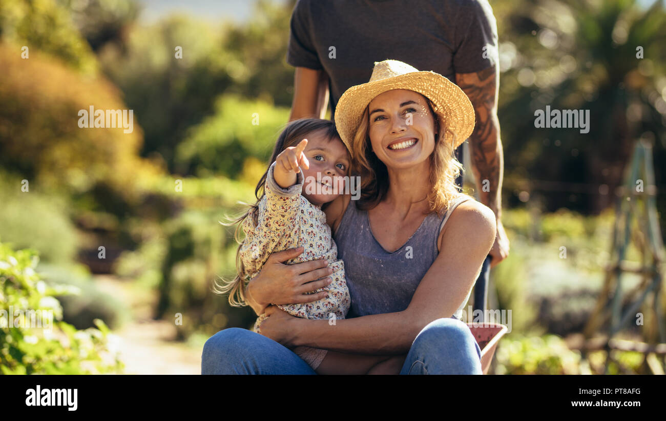 Happy mother and daughter ride en brouette poussée par l'homme dans la ferme. Femme avec petite fille en pointant à l'écart pendant qu'à l'extérieur. brouette Banque D'Images