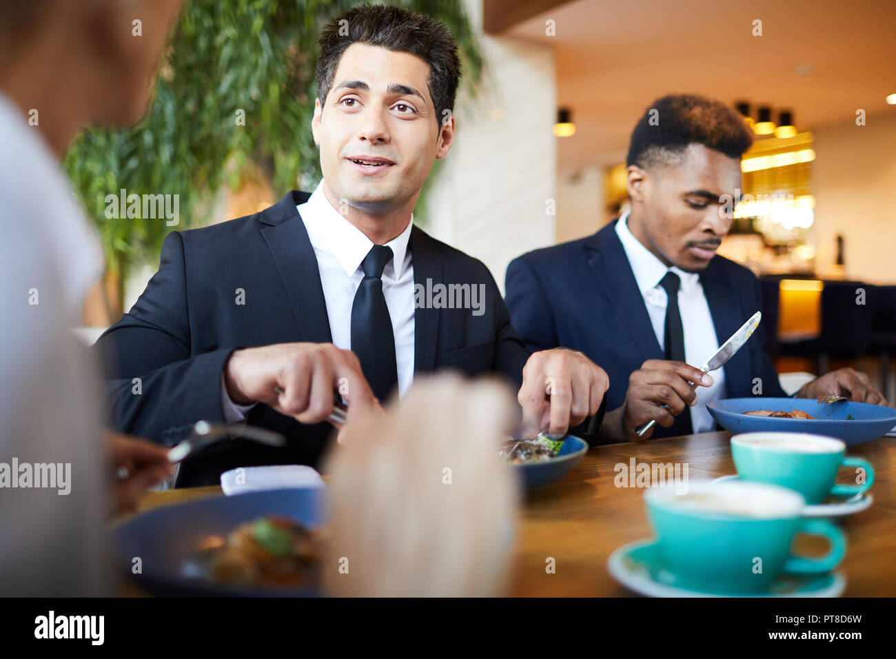 Beau jeune homme d'affaires positif en costume officiel assis à table et de manger tout en discutant avec des collègues de la salade pour le dîner au restaurant Banque D'Images