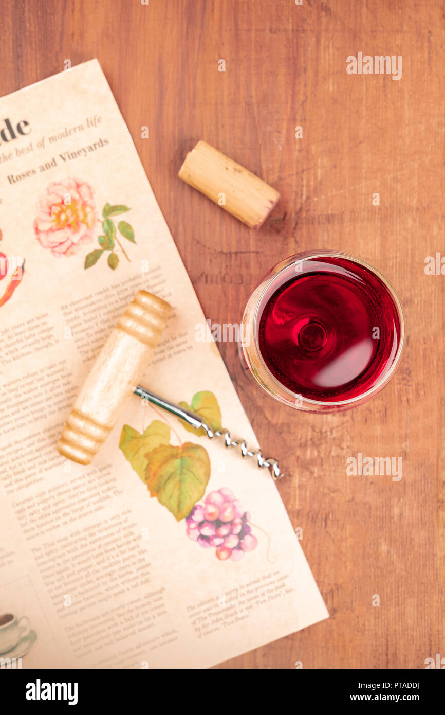 Une photo prise à la verticale d'un verre de vin rouge avec un tire-bouchon et un bouchon vintage, tourné sur un flou vintage journal avec un dessin d'une grappe de raisins sur une vigne Banque D'Images