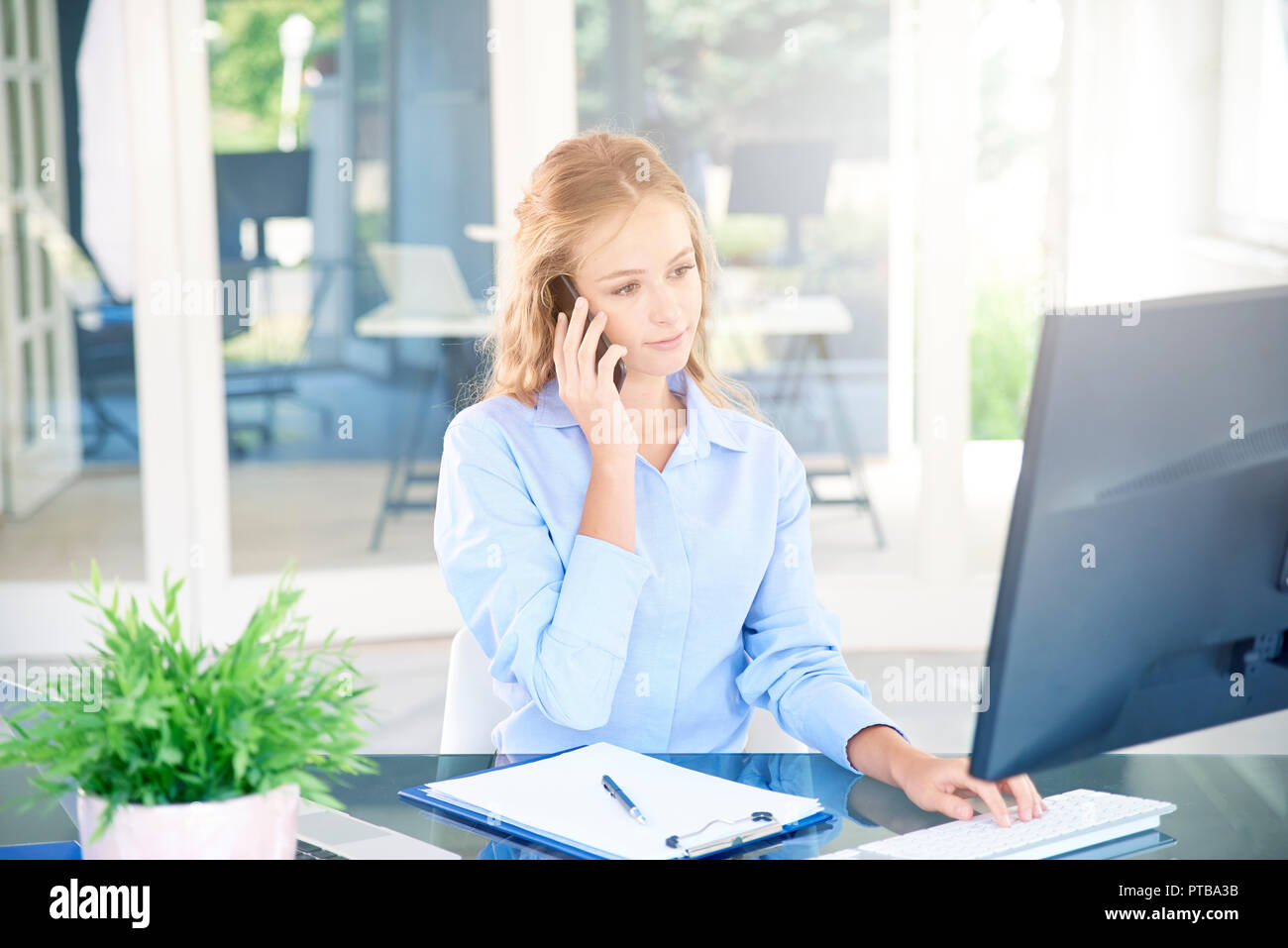 Portrait de belle jeune femme d'adjoint faisant appel alors qu'assis à un bureau et travailler sur ordinateur. Banque D'Images