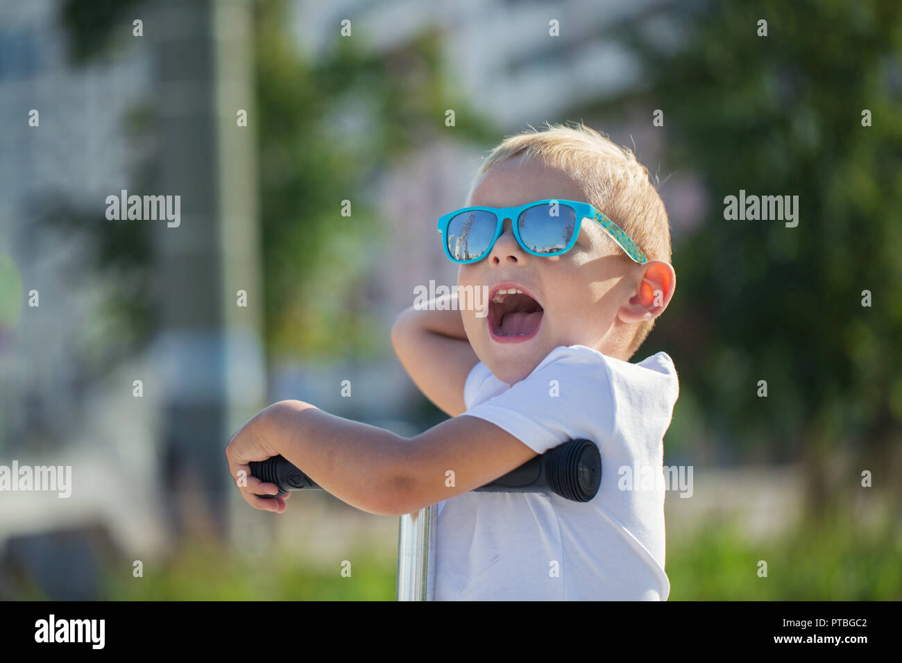 Beau petit Garçon à lunettes élégant monte un scooter sur une piste dans le parc sur une journée ensoleillée Banque D'Images