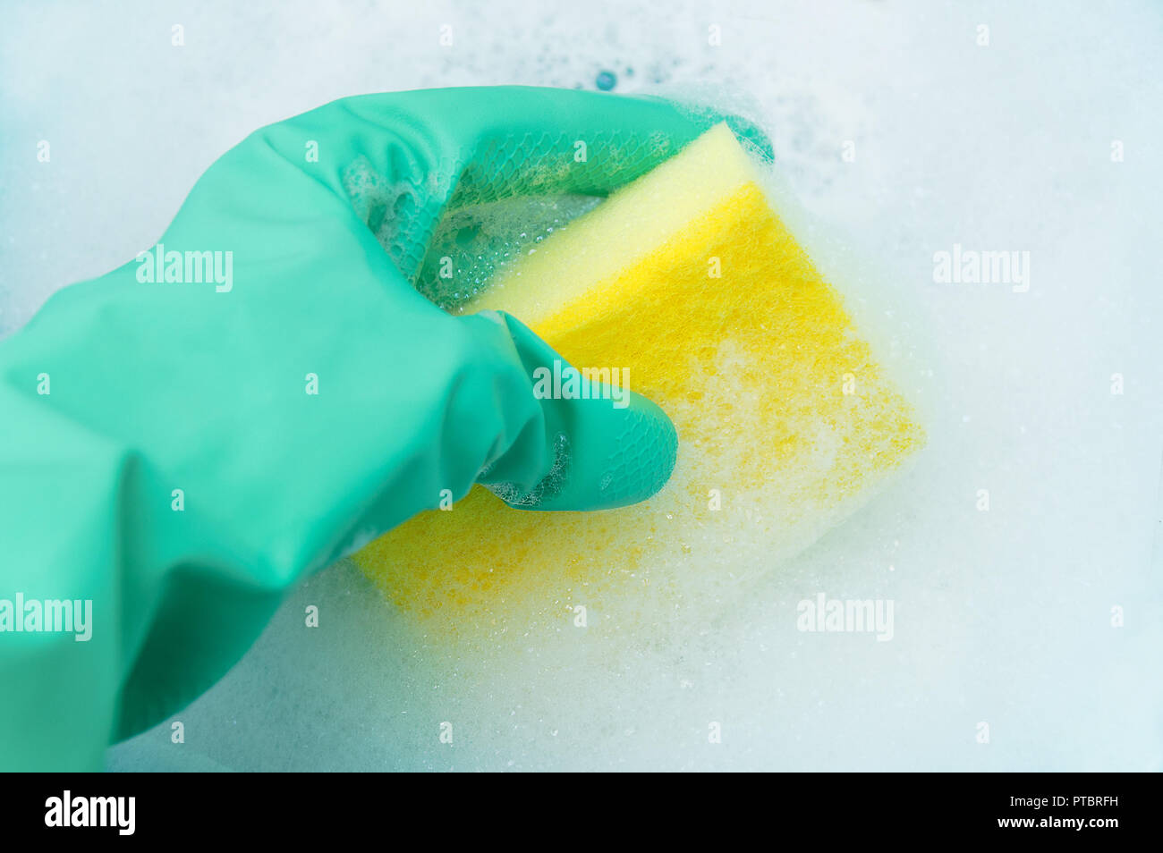 Une main dans un gant turquoise est titulaire d'une éponge pour laver la vaisselle dans une mousse abondante. Close-up Banque D'Images