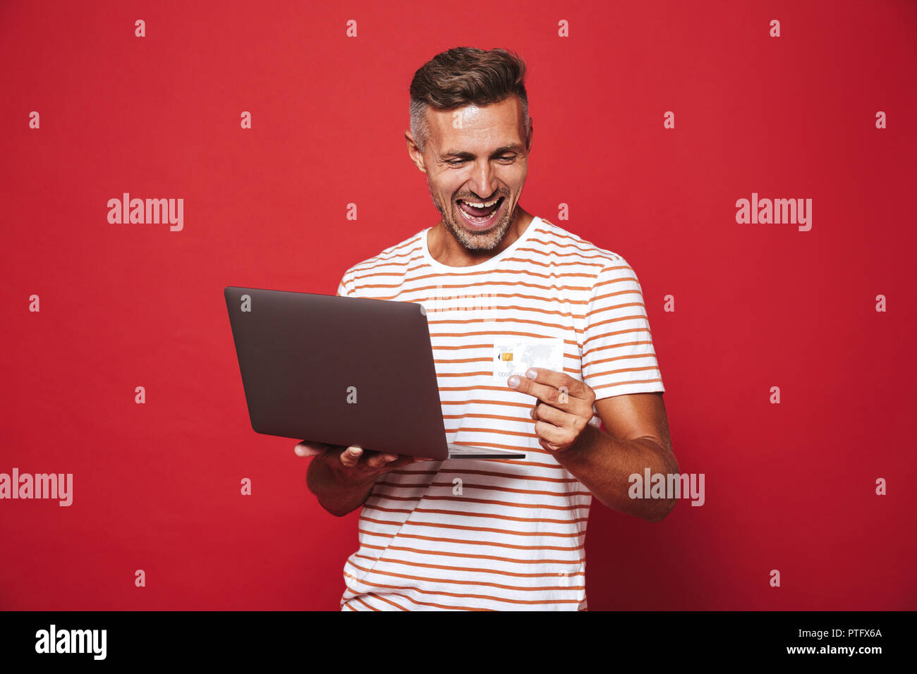 Image de l'homme barbu en t-shirt à rayures smiling while holding credit card et ordinateur portable isolé sur fond rouge Banque D'Images