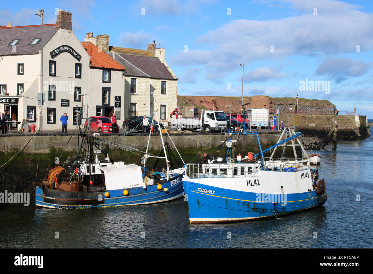 Les gens de port sur quai Eyemouth regarder un petit bateau de pêche entrant dans le port passé un autre bateau déjà attaché au quai. Banque D'Images