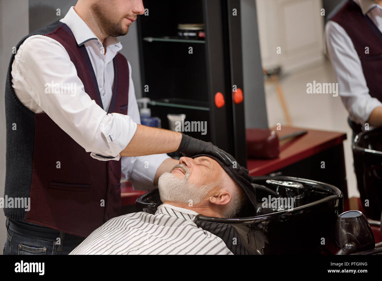 Méconnaissable coiffure tête de balayage avec la clientèle en noir serviette lavabo. Coiffure homme cheveux style de vieux client avec barbe grise. Hairstylist travaillant dans un salon de barbier. Banque D'Images