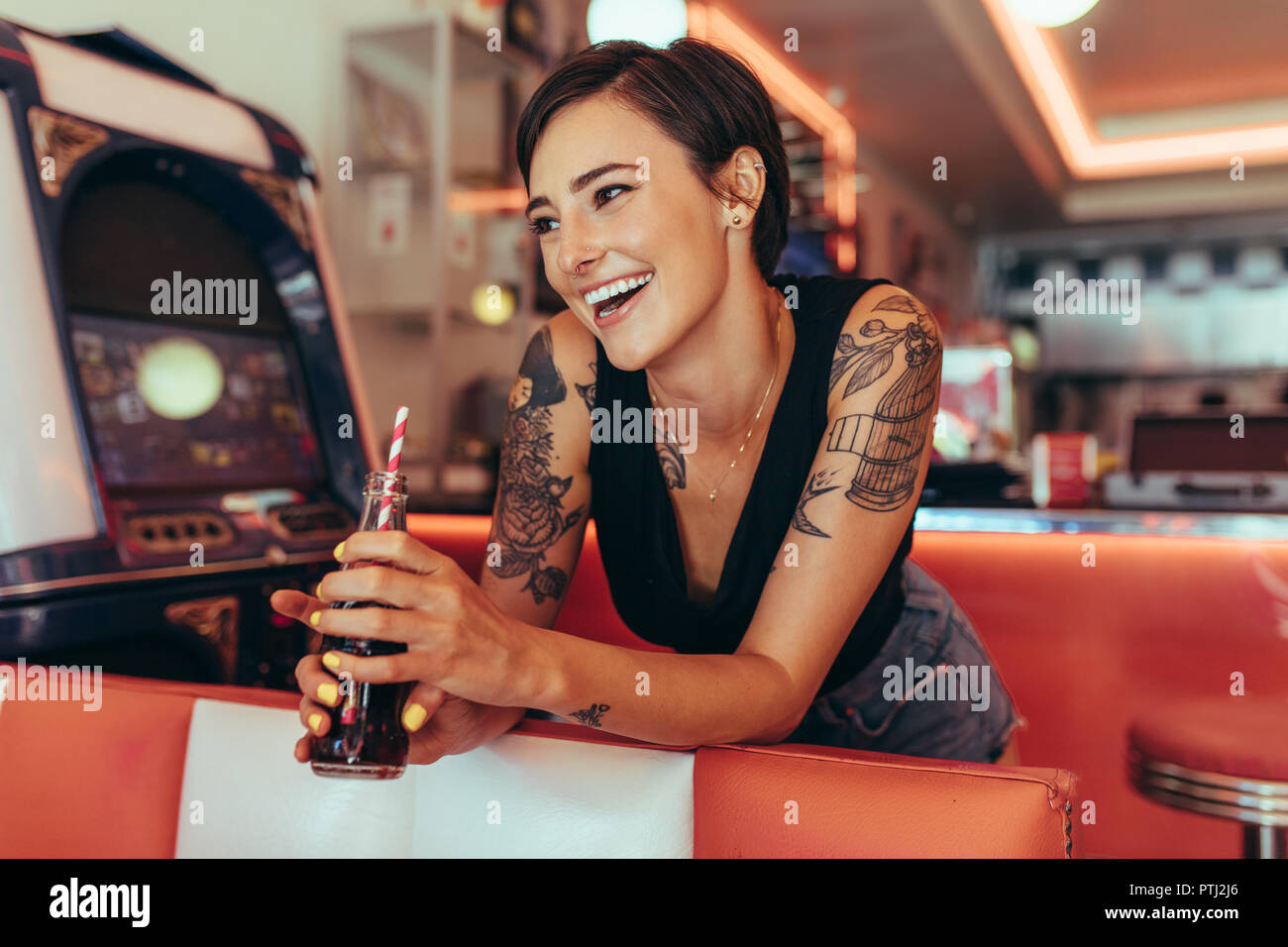 Smiling woman standing in a diner holding a soft drink à l'hôtel. Femme aux bras tatoués boivent des boissons avec une paille dans un restaurant. Banque D'Images