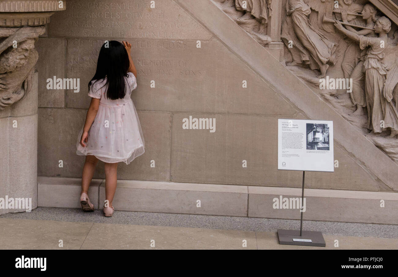 Une petite fille en robe blanche suit le contour des lettres d'une inscription gravée dans le mur dans le Metropolitan Museum. Banque D'Images