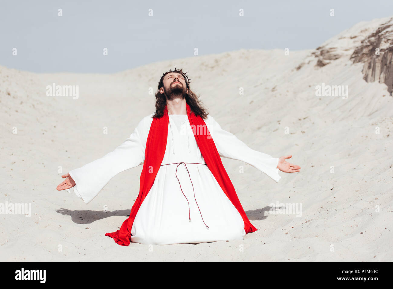 Jésus en robe, ceinture rouge et couronne d'épines debout sur les genoux avec les bras ouverts dans le désert Banque D'Images