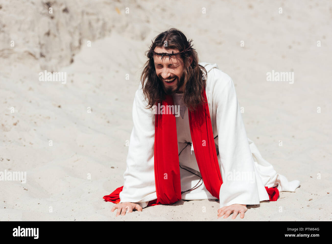 Rire Jésus dans robe, ceinture rouge et couronne d'épines debout sur les genoux et toucher le sable avec les mains dans le désert Banque D'Images