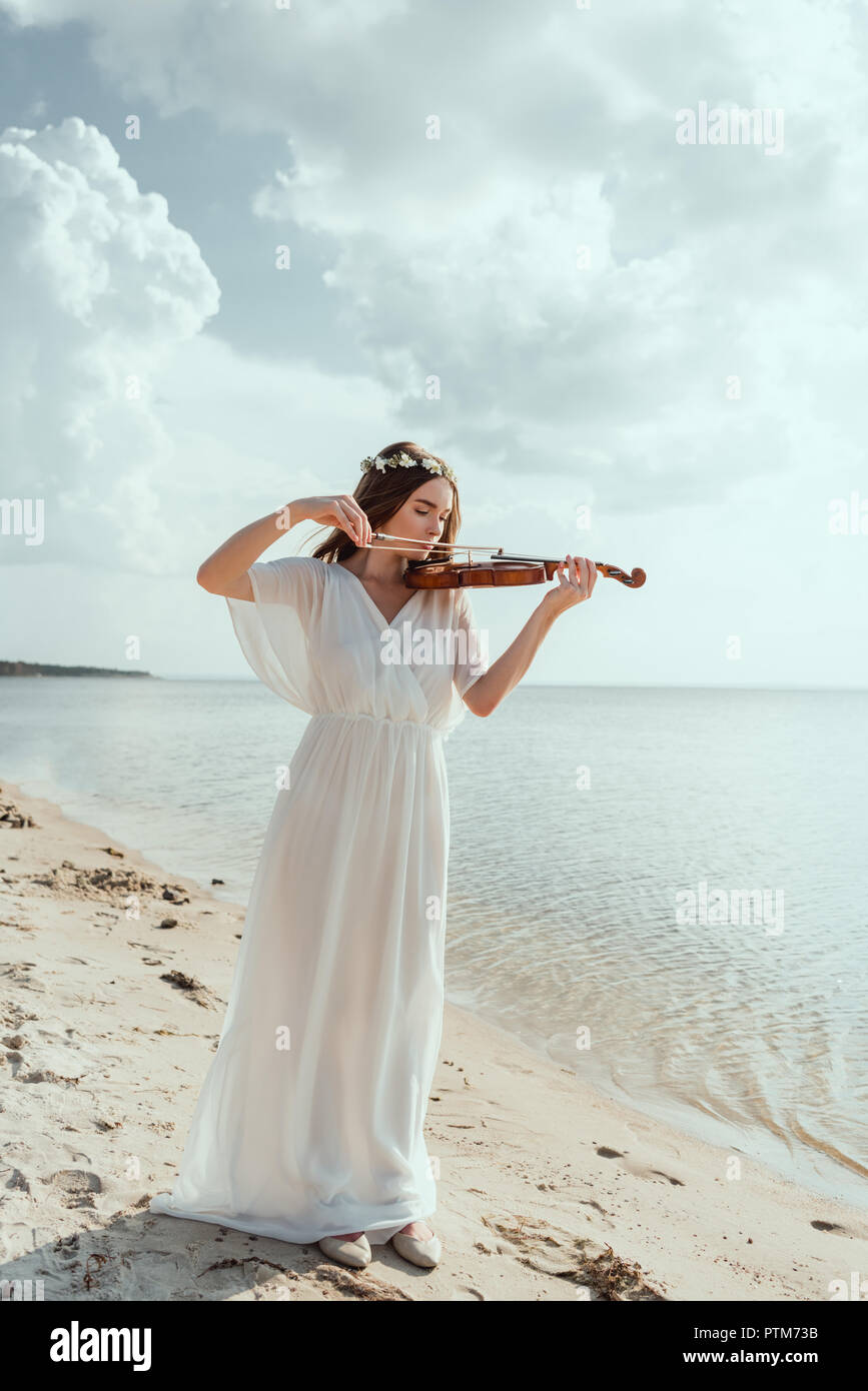 Belle fille élégante en robe blanche et couronne de fleurs à jouer du violon  sur la plage de sable Photo Stock - Alamy