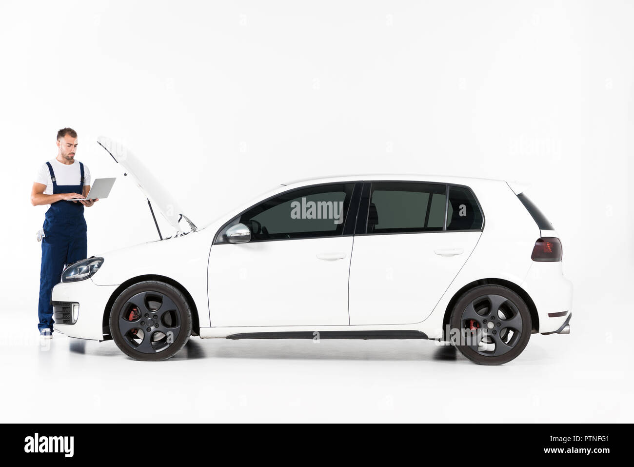 Beau mécanicien d'automobile à l'aide d'ordinateur portable à proximité de capot de voiture sur blanc Banque D'Images