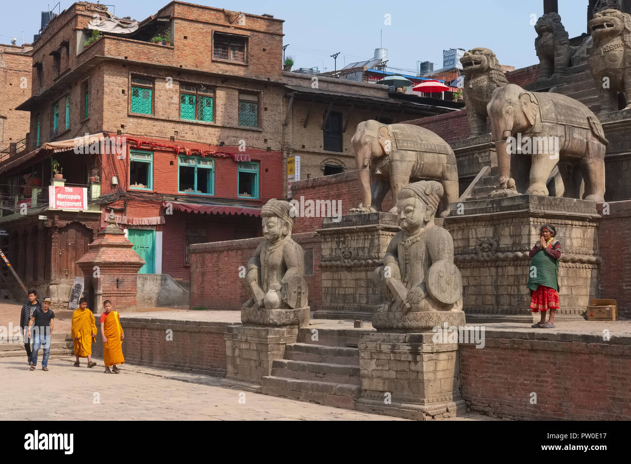 Une femme priant au pied de temple de Nyatapola, flanqué de figures de pierre, Taumadhi Sq., Bhaktapur, Népal, avec deux jeunes moines bouddhistes en passant par Banque D'Images