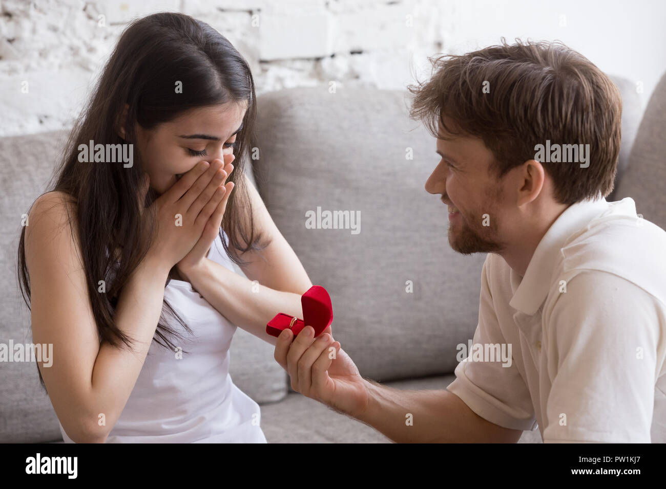 Heureux jeune homme propose à sa copine assise sur canapé Banque D'Images
