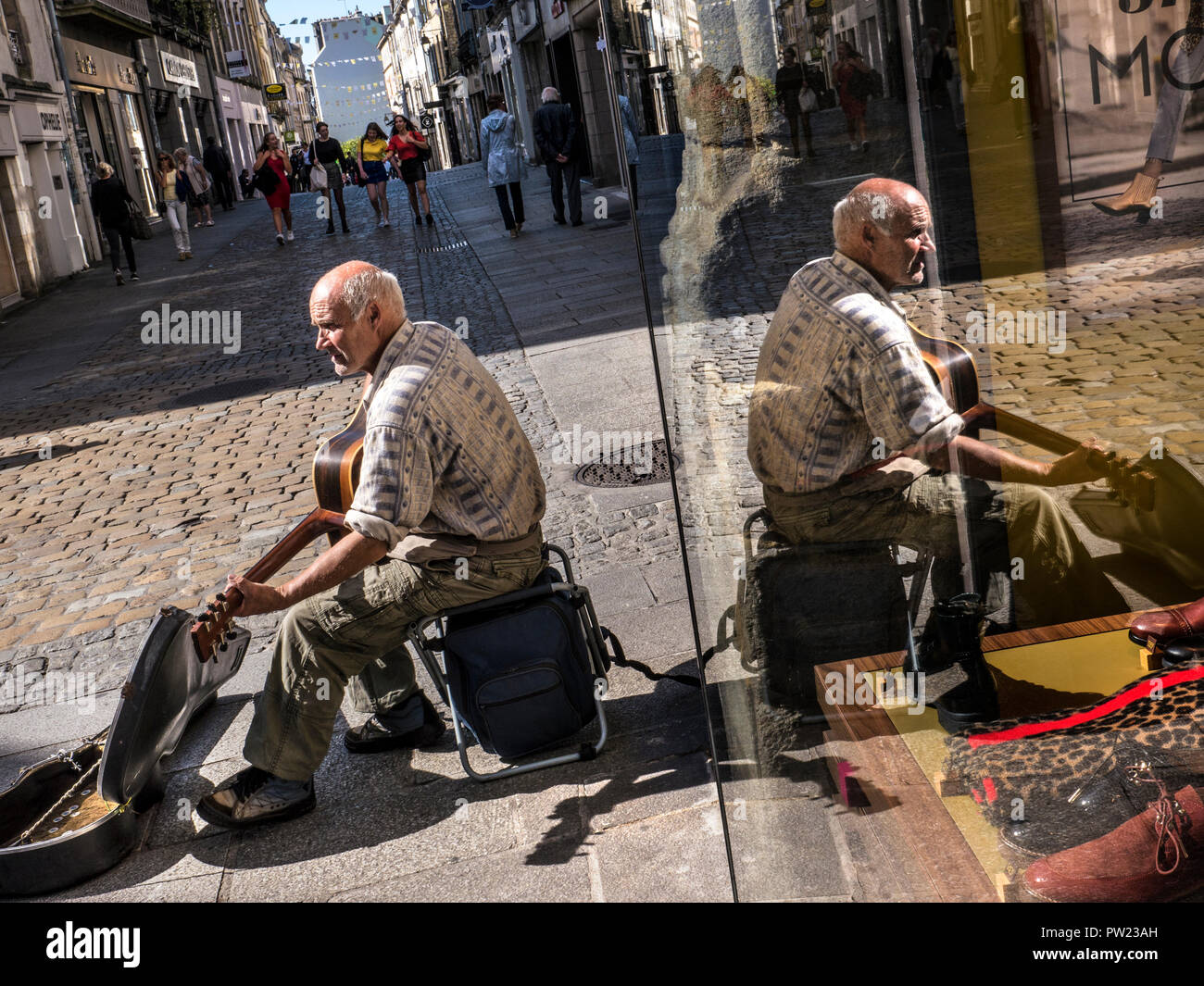 Musicien de rue musicien ambulant guitariste chanteur dans l'arbre de la lumière du soleil, qui se reflète dans la fenêtre que vous recherchiez un magasin de vêtements, dans la vieille ville de Quimper Bretagne France Banque D'Images