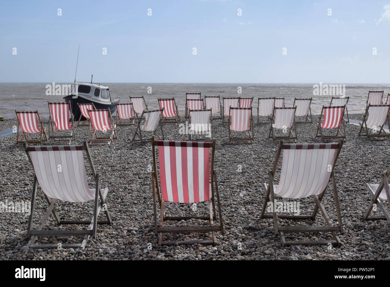 Des rangées de chaises longues rouge et blanc vide sur une plage de galets avec vue sur la mer et le ciel bleu en arrière-plan, petit bateau bleu et blanc sur le côté gauche. Banque D'Images