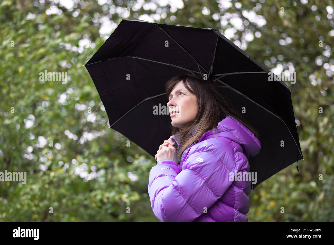Vue latérale rapprochée d'une femme portant un manteau isolée à l'extérieur, tenant un parapluie noir et regardant vers le haut. Banque D'Images