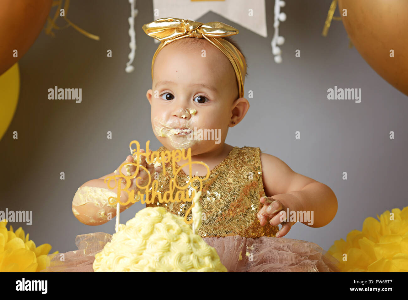 Petite fille Bébé doux dans une robe d'or avec un arc sur la tête d'essayer un gâteau de gelée jazzy une crème. studio shot of un anniversaire sur fond gris entouré par des boules Banque D'Images