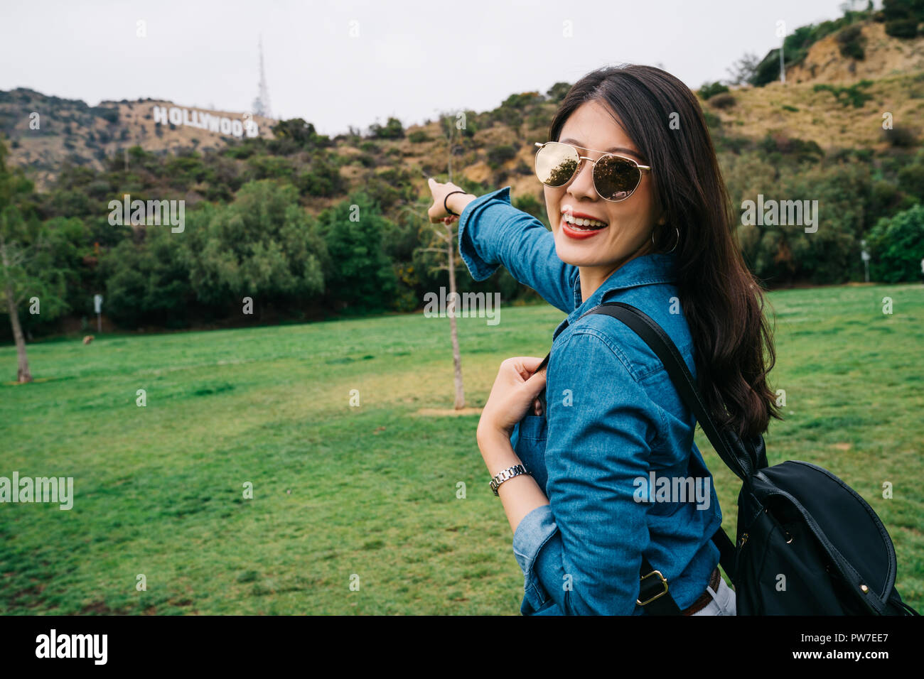 Femme joyeuse, pointant sur l'enseigne Hollywood, face à la caméra en souriant. dame en voyage de vacances de printemps à lunettes. Profitant du soleil et vert Banque D'Images