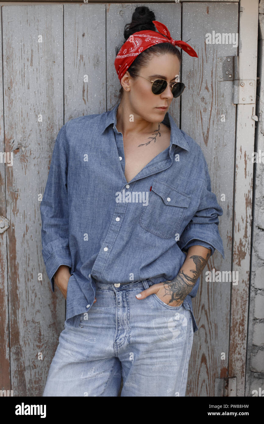La mode femme Cool hipster avec des lunettes avec un tatouage sur les mains d'un denim shirt et jeans vintage avec un fichu rouge se tient près d'un mur en bois Banque D'Images