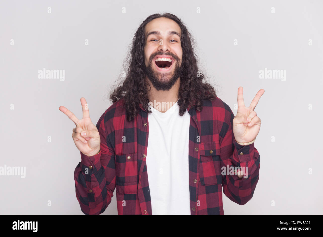 Heureux bel homme avec barbe et cheveux longs noirs dans un style décontracté, chemise rouge à carreaux debout avec la victoire ou signe de paix et looking at camera Banque D'Images Heureux bel homme avec barbe et cheveux longs noirs dans un style décontracté, chemise rouge à carreaux debout avec la victoire ou signe de paix et looking at camera Banque D'Images