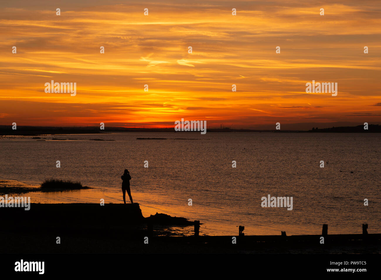 Une femme se tient sur un brise-lames, admirant le coucher du soleil à Seasalter, Whitstable, Kent, Royaume-Uni. Elle regarde à travers l'estuaire de Swale vers l'île de Sheppey. Banque D'Images