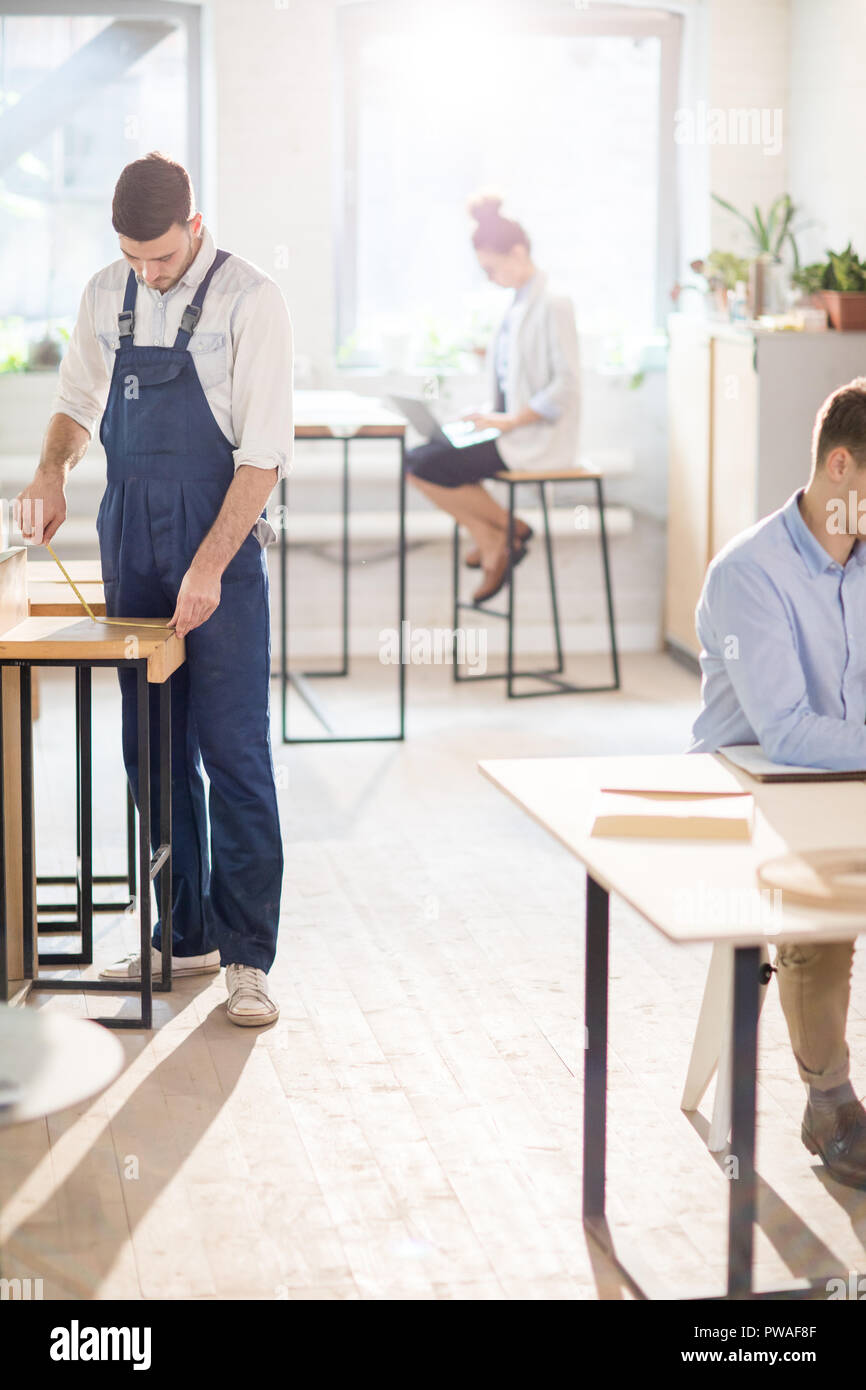 Jeune ingénieur en salopette measuring wooden table pendant que ses deux collègues qui travaillent par un bureau Banque D'Images
