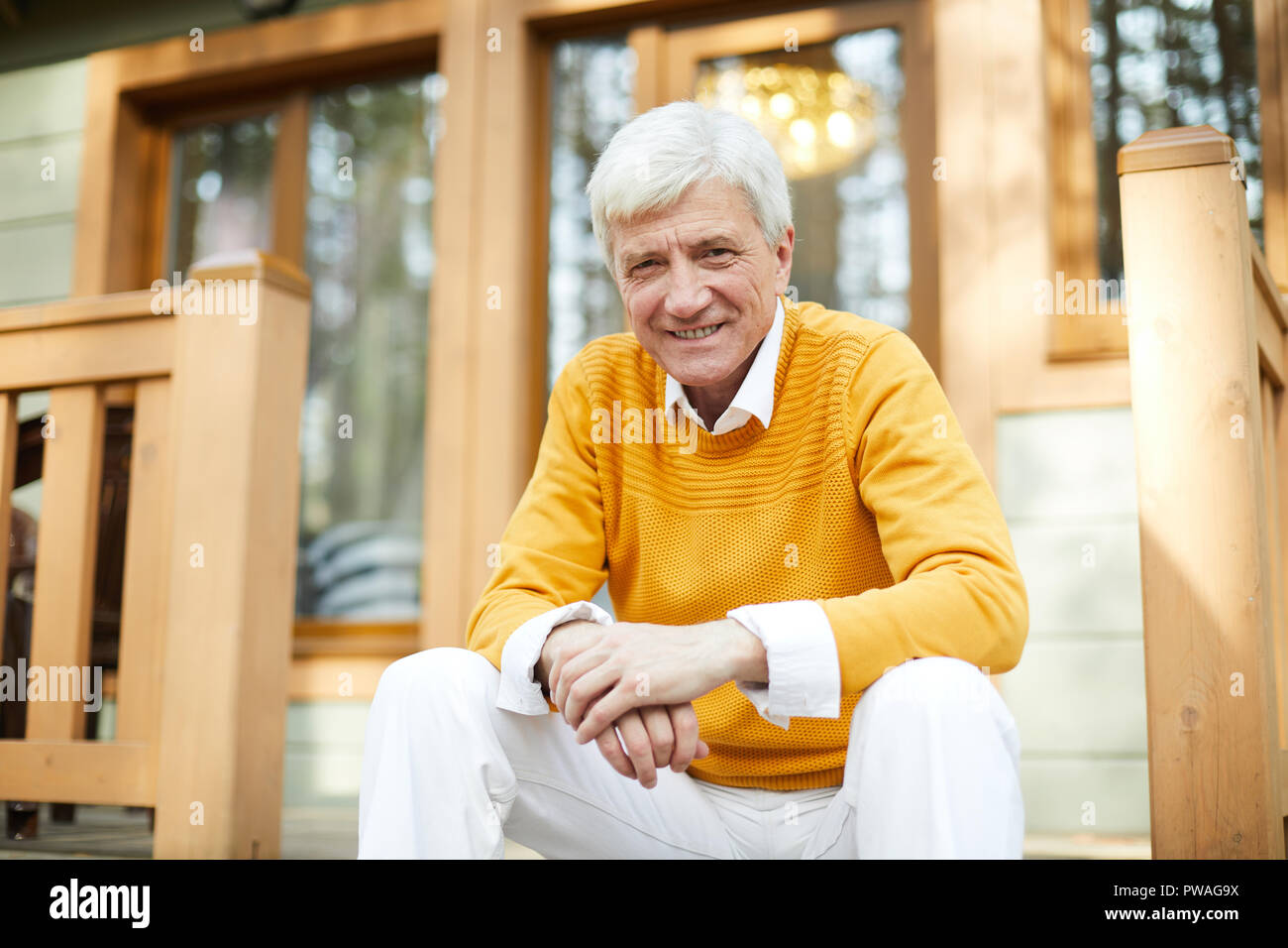 Heureux homme mûr dans casualwear assis sur la terrasse en bois de sa maison de campagne et vous regarde Banque D'Images