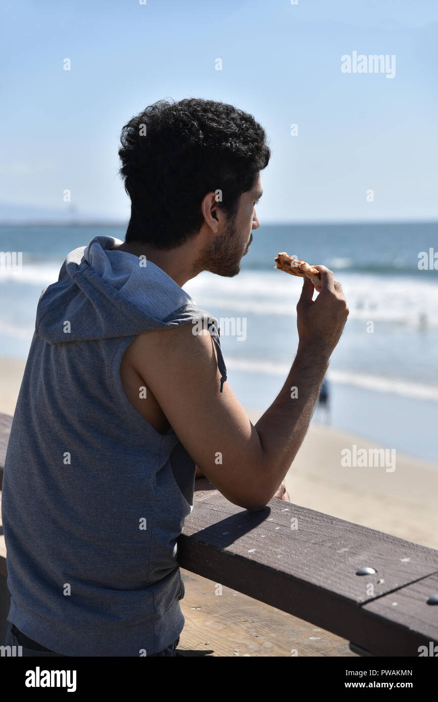 Beau jeune homme mangeant une tranche de pizza sur le quai donnant sur l'océan Pacifique Banque D'Images